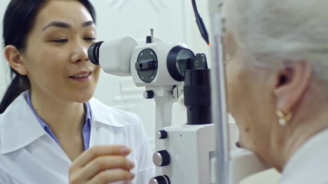 Professional female optometrist looking through binocular of slit lamp and talking to senior woman during eye exam in clinic