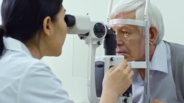 Panning shot of female ophthalmologist doing eye exam with slit lamp while checking eyesight of elderly man