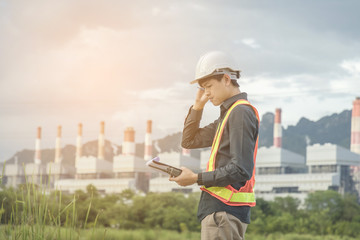 An engineer wearing safety gear inspects an industrial plant using a tablet device while outdoors. The image conveys themes of technology, safety, and industrial operations.