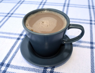 A cup of coffee with milk on a white tablecloth. Cup and saucer in gray with a shade of blue. View from above.