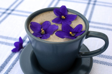 A cup of coffee with milk on a white tablecloth. Cup and saucer in gray with a shade of blue. View from above. The composition is decorated with violet flowers.