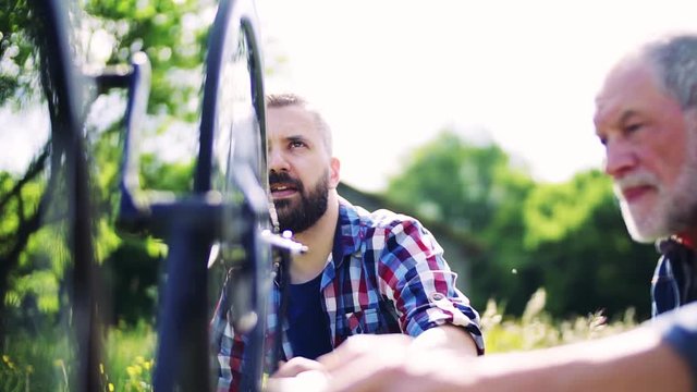 An adult hipster son and senior father repairing bicycle outside on a sunny day.