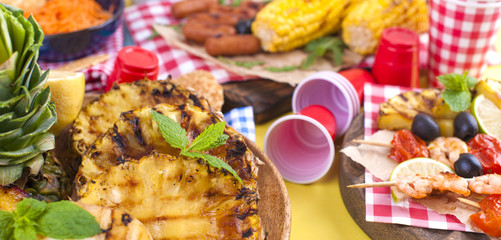 Picnic with grilled food. Sausages and corn on barbecue, shrimp, vegetables and fruits. Delicious summer lunch and plastic dishes. Top view