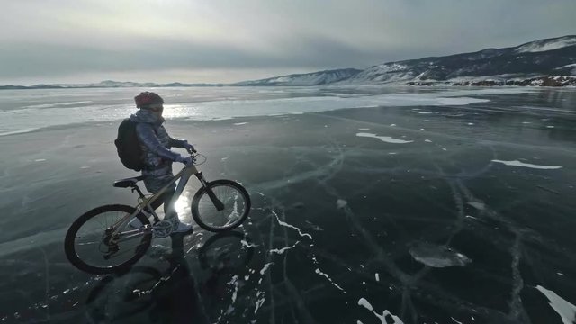 Woman is walking beside bicycle on the ice. The girl is dressed in a silvery down jacket, backpack and helmet. Ice of the frozen Lake Baikal. The tires on the bicycle are covered with special spikes