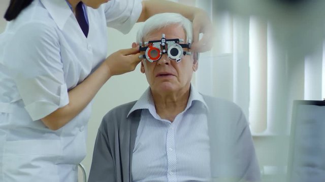 Tracking shot of senior man trying on lenses in trial frame with help of female eye doctor in ophthalmology clinic