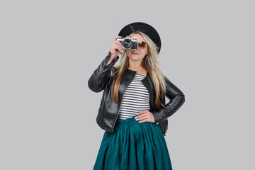 Fashion look, happy cool young woman model with retro film camera makes a photo. wearing an elegant hat, black jacket, blondy hair outdoors over grey background