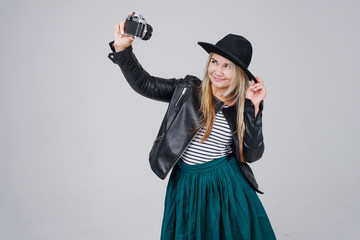 Fashion look, pretty cool young woman model with retro film camera make selfie. wearing an elegant hat, black jacket, blondy hair outdoors over grey background