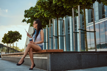 Happy beautiful brunette girl with tattoo on her leg wearing trendy clothes and sunglasses holds a takeaway coffee and a digital tablet while sitting on bench against a skyscraper, looking away