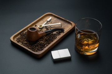 a smoking pipe with pipe tamper tool and tobacco in wooden tray, a glass of whisky and chrome lighter on the black table