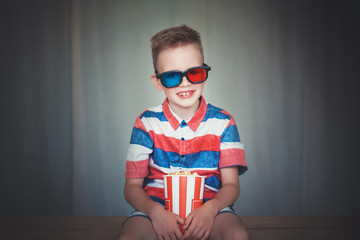 Naklejka premium Young boy watch a movie in 3D glasses at the cinema or at home. Little kid eat popcorn over gray background. Home theater. Cute Child in vintage cinema eyeglasses