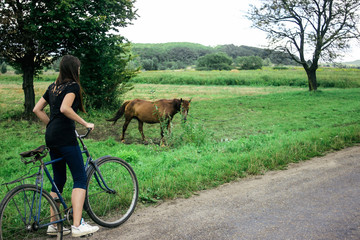 stylish brunette girl on old bicycle looking at brown horse in country site, summer travel