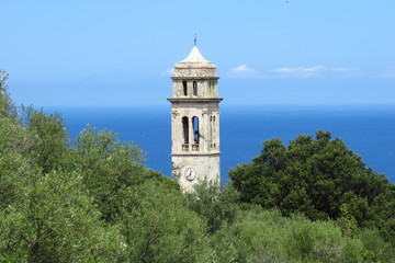 Obraz premium The church tower of Pino surrounded by lush green trees against a blue sea and sky background, Haute-Corse, Corsica, France