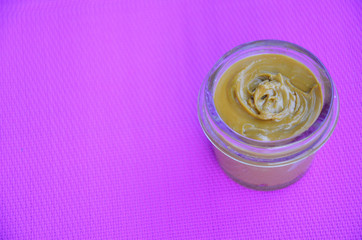 Jar and spoon of peanut butter and peanuts on purple background from top view. copy space