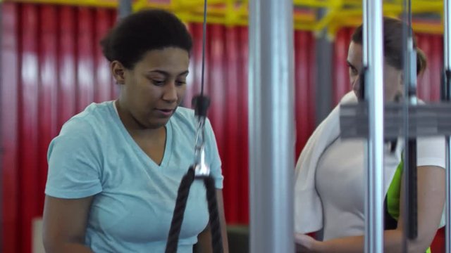 Medium shot of focused black woman doing triceps pushdown exercise as her female friend with towel giving her advice on posture