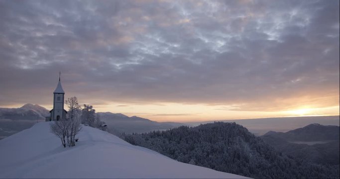 Winter sunrise time lapse of a snowy landscape of the popular and historic Slovenian church of St. Primus and Felician on Jamnik hill. Alps can be seen in the background with clouds moving above.
