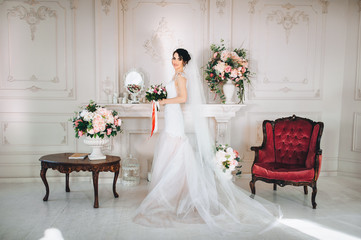 A beautiful bride stands in a white studio, against a background of flowers, candles and a red chair. Wedding portrait of a young bride.