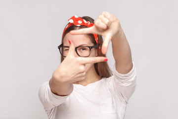 Fototapeta premium choosing composition or focusing portrait of beautiful emotional young woman in white t-shirt with freckles, glasses, red lips and head band. indoor studio shot, isolated on light gray background.