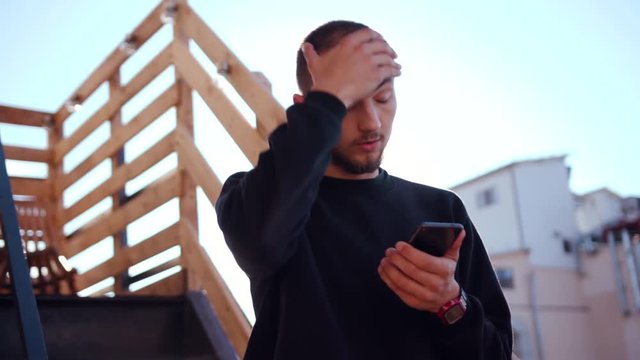 Handsome baffled young man holding smartphone in hand, touching head. Portrait of good-looking upset student wearing casual clothes. Bad news.