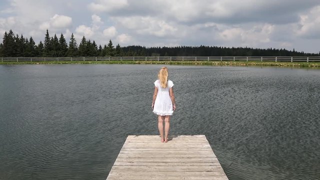Lonely blonde long hair woman standing on wooden pier at the windy summer lake.