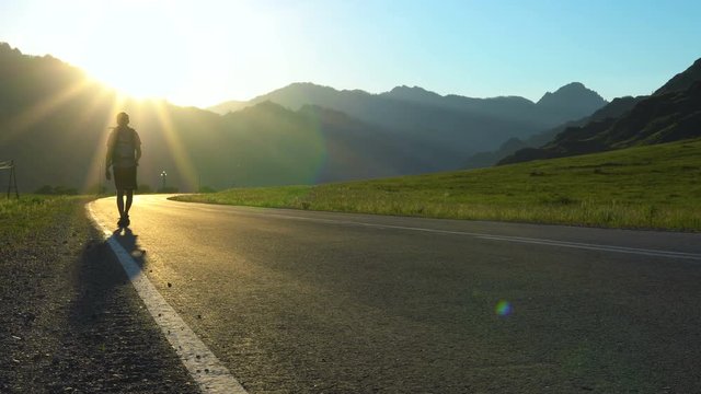 A lone young tourist with a backpack walks along an empty asphalt road in a mountainous area. It's sunset time.