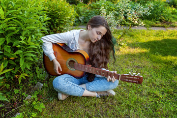 Young hipster woman sitting in grass and playing guitar on park or garden background. Teen girl...