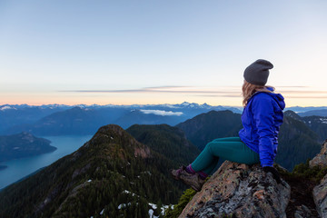 Naklejka premium Adventurous woman on top of a mountain cliff is enjoying the beautiful summer sunrise. Taken on Mount Brunswick, Lions Bay, North of Vancouver, BC, Canada.