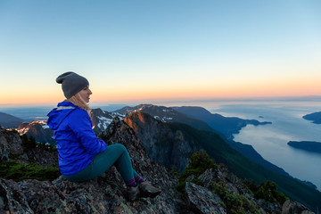 Adventurous woman on top of a mountain cliff is enjoying the beautiful summer sunrise. Taken on Mount Brunswick, Lions Bay, North of Vancouver, BC, Canada.