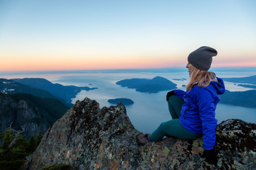 Adventurous woman on top of a mountain cliff is enjoying the beautiful summer sunrise. Taken on Mount Brunswick, Lions Bay, North of Vancouver, BC, Canada.