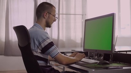 Cute Caucasian guy in glasses sitting at computer, typing on keyboard. Glad young man showing something in monitor, looking at camera and giving thumbs up.
