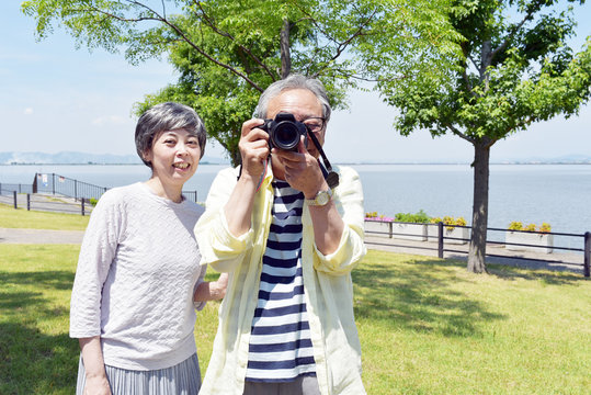 海の近くの公園で、笑顔でカメラを構えるシニア夫婦