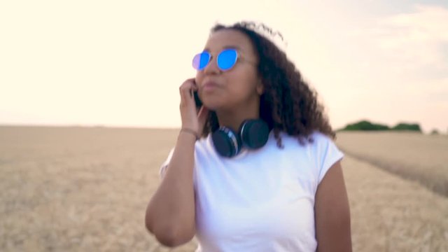 Slow motion follow shot video of beautiful mixed race African American girl teenager young woman in white T-shirt and blue sunglasses walking and talking on her cell phone