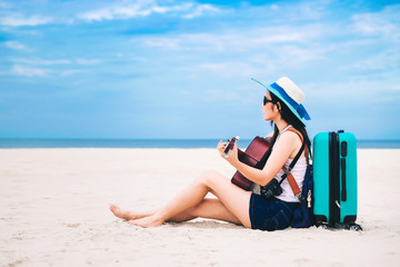 Woman traveler sitting on a baggage and playing a guitar on the beach during holiday.