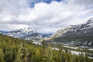Wide angle mountain view with cloudy skies, snow covered peaks and valley of spruce trees.