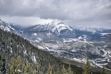 Wide angle mountain view with cloudy skies, snow covered peaks and valley of spruce trees.