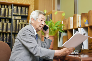 重役室で、書類を片手にスマホで電話をするシニアビジネスマン