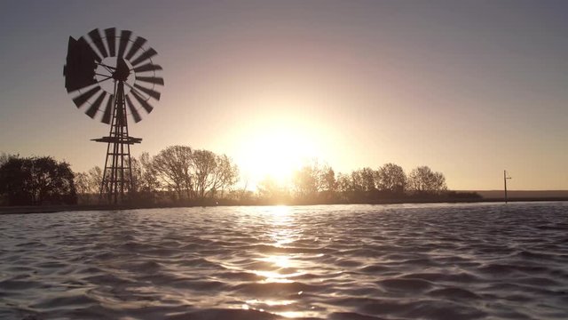Water ripples in farm reservoir as vintage windmill water pump turns in the wind at sundown.