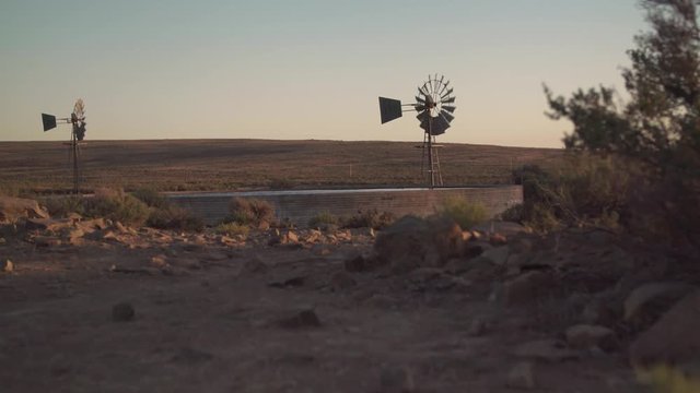 Two old fashioned farm windmills on dry arid land spin in the wind.