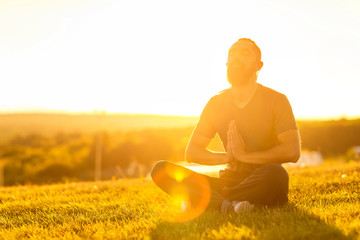 Bearded man is meditating in lotus yoga pose at summer outdoor sunset background sitting at a...