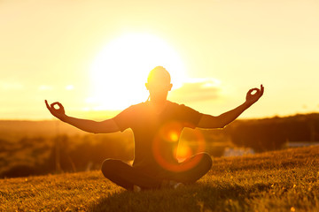Silhouette of bearded man meditating in lotus yoga pose at summer outdoor sunset background sitting at a golden field.
