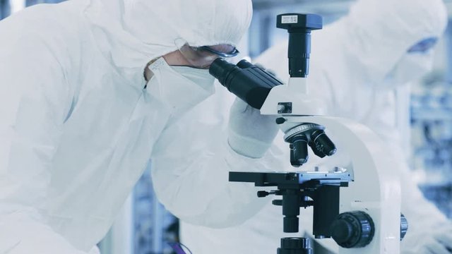 In Laboratory Scientist in Protective Clothes Doing Research, Using Microscope and Writing Down Data