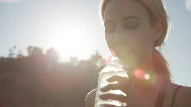 Beautiful blonde girl drinks from water bottle outside with the sun shining and blue sky