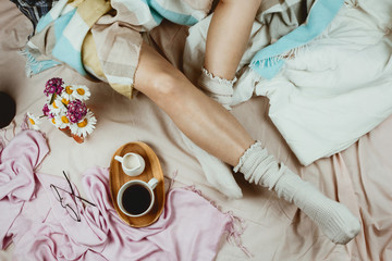 Cozy flatlay with white tanned woman in white socks sitting in her bed with cup of black coffee and...