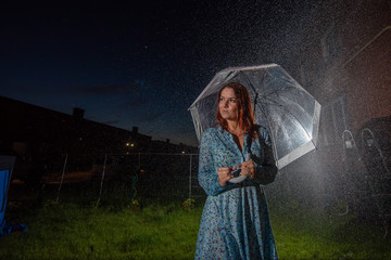 Sad young woman in a dress in the rain with a transparent umbrella at night. Beautiful woman with a...