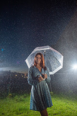 Sad young woman in a dress in the rain with a transparent umbrella at night. Beautiful woman with a transparent umbrella in the lanterns and rain drop