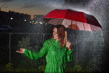 Young woman in green raincoat in rain with red umbrella at night. Smiling young woman in the rain...