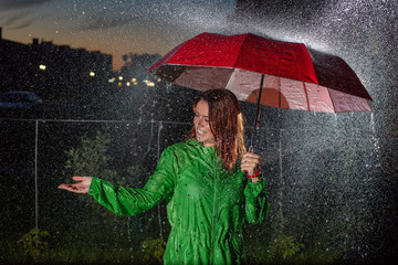 Young woman in green raincoat in rain with red umbrella at night. Smiling young woman in the rain with an umbrella in the evening. Beautiful woman with red umbrella in lanterns and rain drops