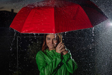 Young woman in green raincoat in rain with red umbrella at night. Smiling young woman in the rain...