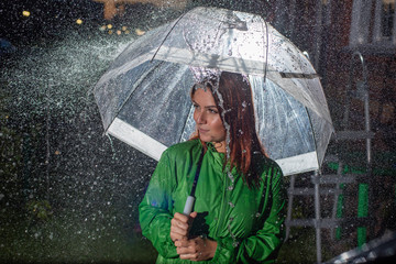 Smiling young woman in the green coat in the rain with a transparent umbrella in the night.  Beautiful woman with a transparent umbrella in the lanterns and rain drop