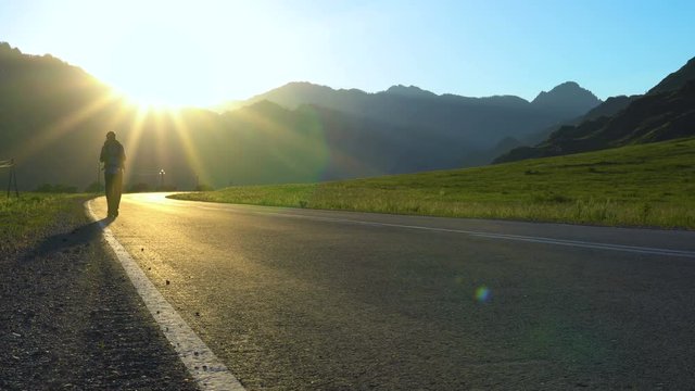 A lone young tourist with a backpack walks along an empty asphalt road in a mountainous area. It's sunset time.