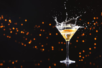Glass with splashing cocktail and slice of orange against blurred lights